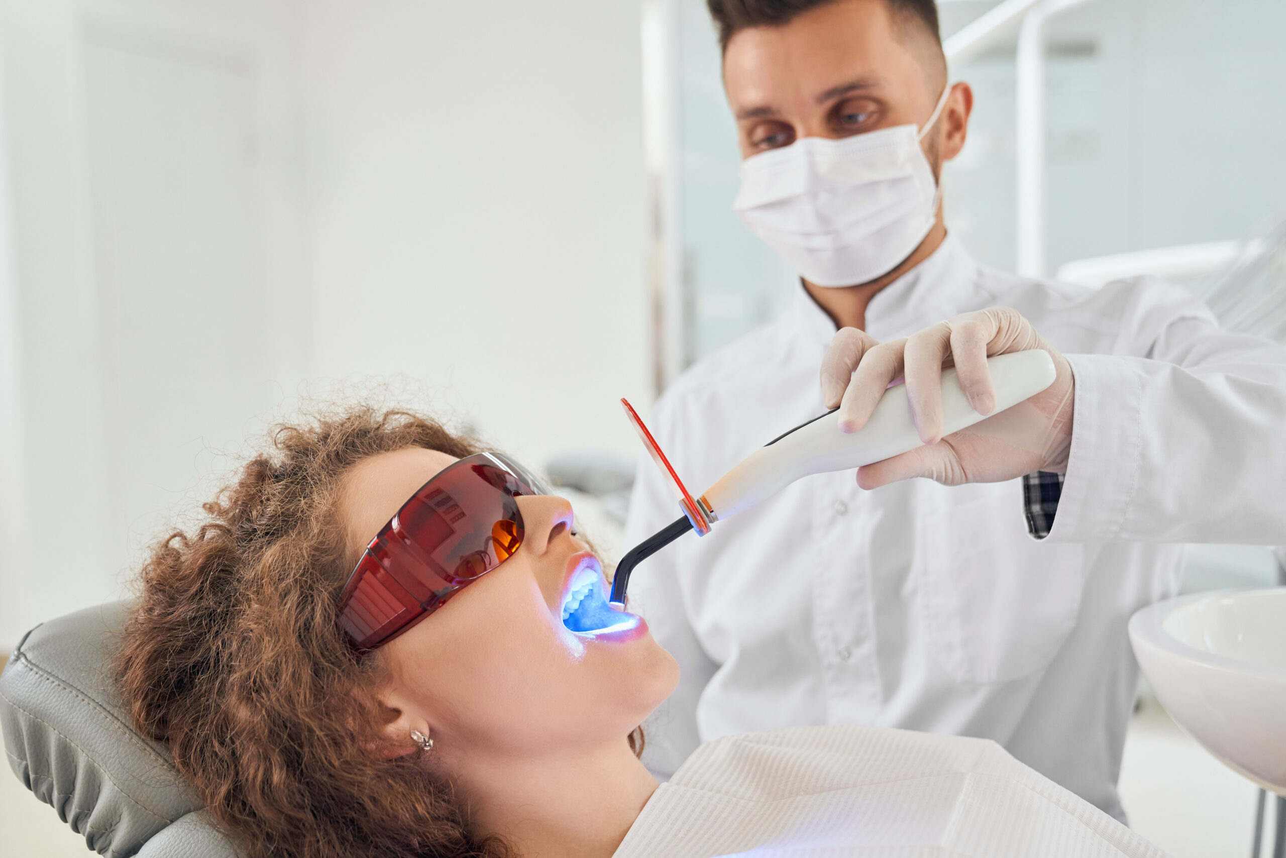 Side view of attractive female patient in protective glasses lying on dental chair with opened mouth while male dentist in white uniform and mask keeping ultraviolet in clinic. Concept of medicine.